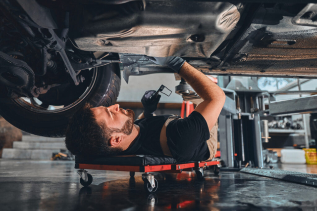 Male car mechanic worker working using wrench tool for repair, maintenance underneath car. Mechanic vehicle service checking under car in garage. Services